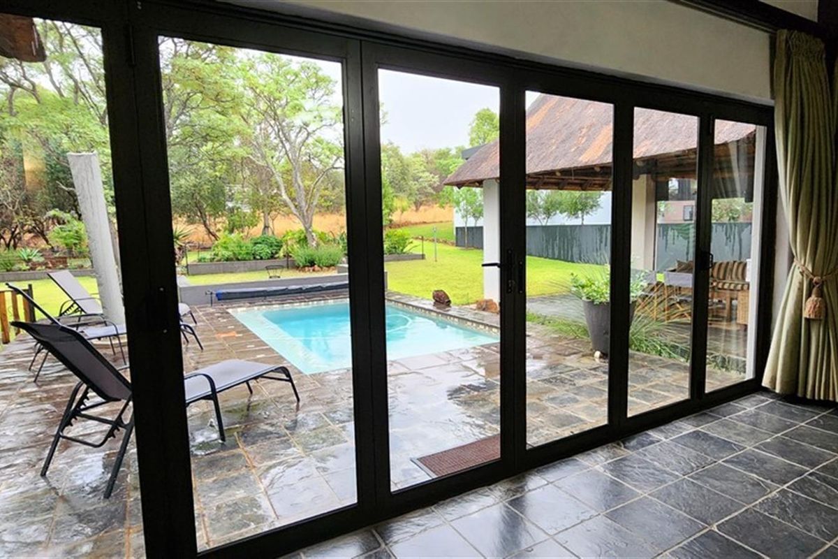 A view from an interior living space through large black-framed folding doors onto a paved patio area with a swimming pool and garden.