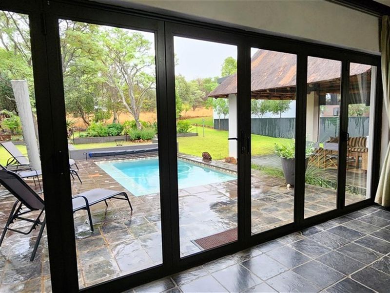 A view from an interior living space through large black-framed folding doors onto a paved patio area with a swimming pool and garden.