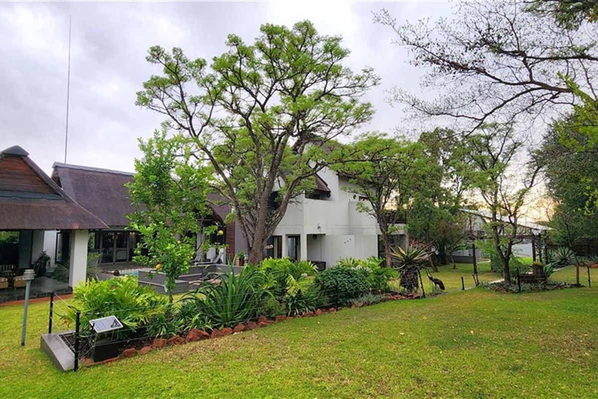 A view of the rear exterior of the house, featuring a thatched roof patio area, landscaped garden, and mature trees.