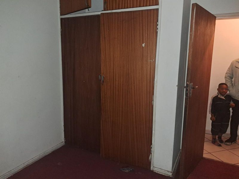 A bedroom featuring built-in wooden cupboards and carpeted flooring, showing signs of wear and requiring cosmetic updates.