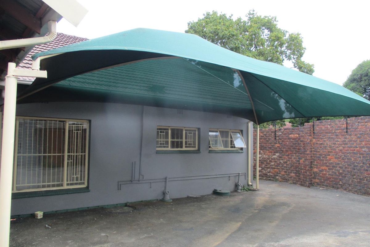 Rear exterior area featuring a large green shade sail, brick boundary wall, and paved driveway space with security-fitted windows.