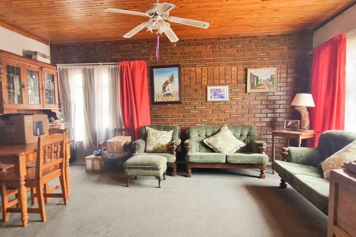 A spacious living room featuring a prominent exposed brick feature wall, timber-paneled ceiling with a central fan, and large windows allowing natural light.