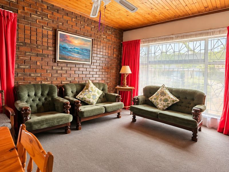 A living room featuring an exposed brick feature wall, timber ceiling, and large windows with security bars, offering a dated but functional living space.