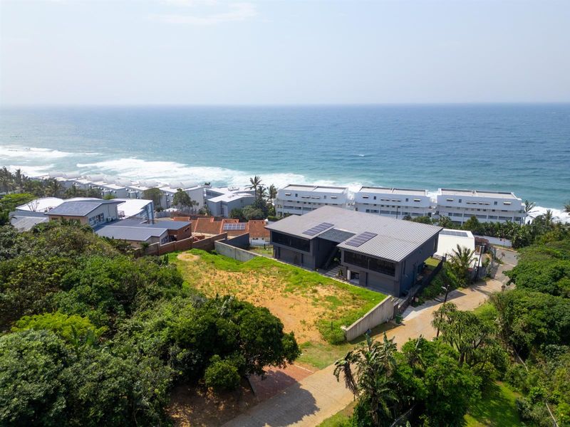 Aerial view of a residential property featuring a modern home with solar panels, situated near the coastline with surrounding greenery and paved access.