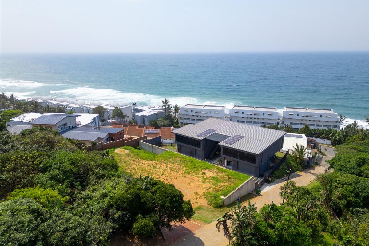Aerial view of a residential property featuring a modern home with solar panels, situated near the coastline with surrounding greenery and paved access.