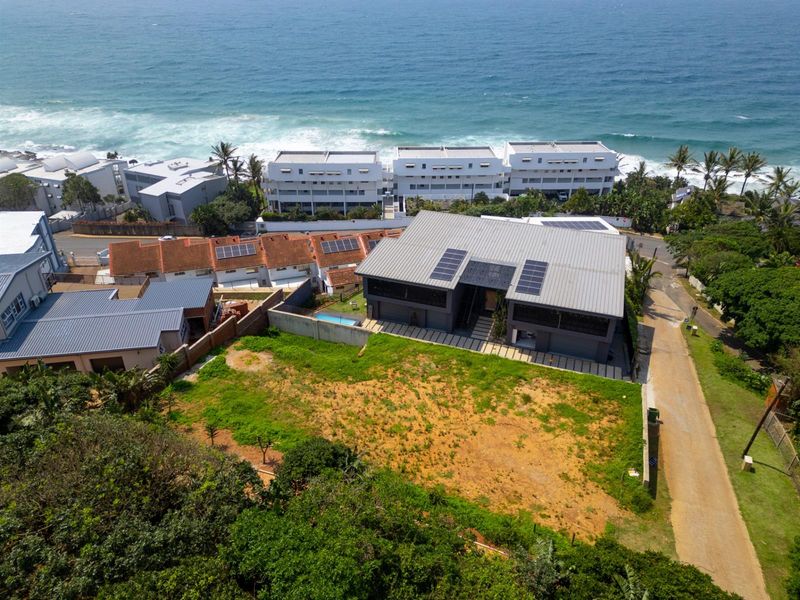 Aerial view of a residential property featuring a modern home with solar panels, situated near the coastline with surrounding neighborhood infrastructure.