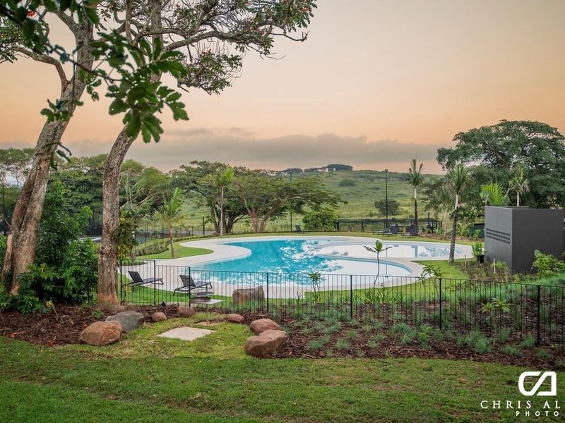 A well-maintained swimming pool area featuring landscaped gardens, paved surrounds, and metal perimeter fencing, set against a scenic natural backdrop.