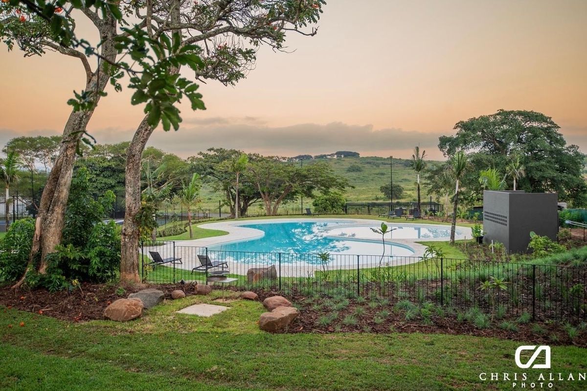 A well-maintained swimming pool area featuring landscaped gardens, paved surrounds, and metal perimeter fencing, set against a scenic natural backdrop.