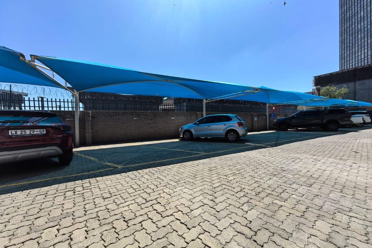 Commercial parking area featuring paved ground surface and blue shade cloth structures for vehicle protection, enclosed by a brick perimeter wall with security fencing.