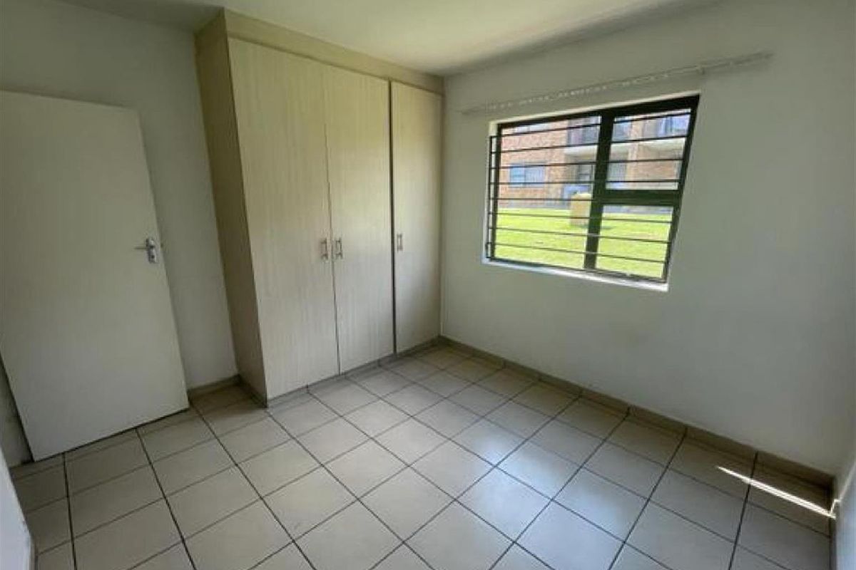 A bright, empty bedroom featuring neutral floor tiles, built-in wooden cabinetry, and a window with security bars overlooking the complex grounds.