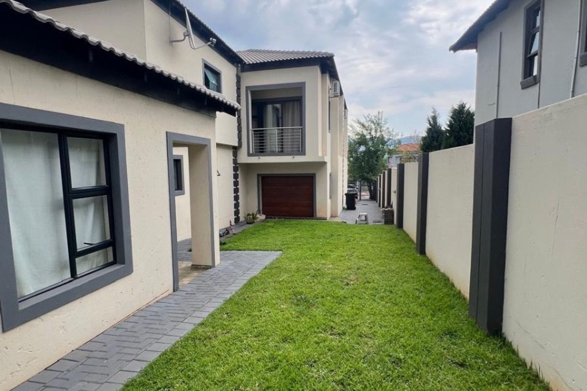 A well-maintained rear garden area featuring a paved walkway, manicured lawn, and a secure boundary wall, with views of the house exterior and balcony.