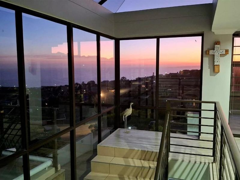 An enclosed balcony or sunroom featuring floor-to-ceiling glass windows, tiled flooring, and a metal balustrade, offering expansive sunset and coastal views.