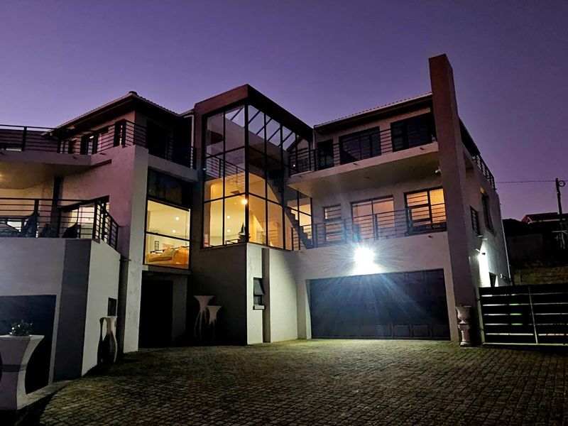 A modern multi-story residence featuring a striking glass facade, multiple balconies with metal railings, and a paved driveway leading to a double garage.
