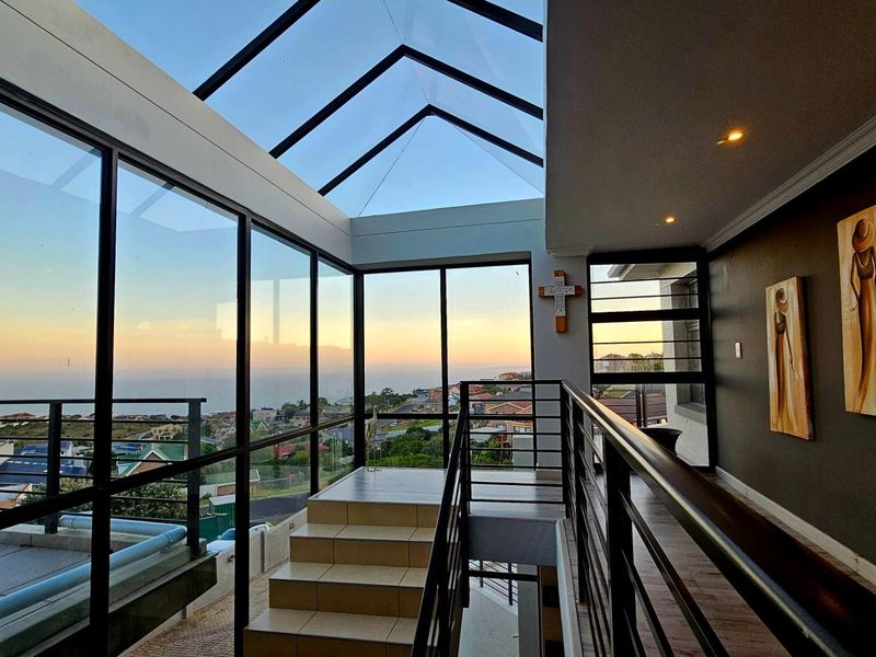 A modern, light-filled enclosed balcony or sunroom featuring a glass roof, metal railings, and expansive views of the surrounding neighborhood and coastline.