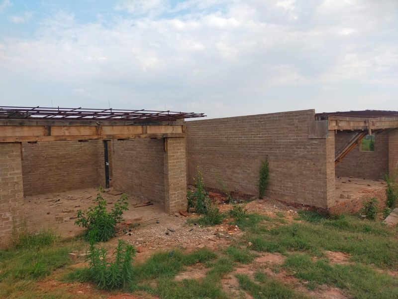 An unfinished residential structure showing exposed brick walls and timber roof framing, currently in the early stages of construction.