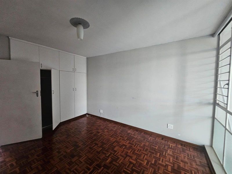 An empty bedroom featuring classic parquet flooring, built-in storage cupboards, and a large window allowing natural light.