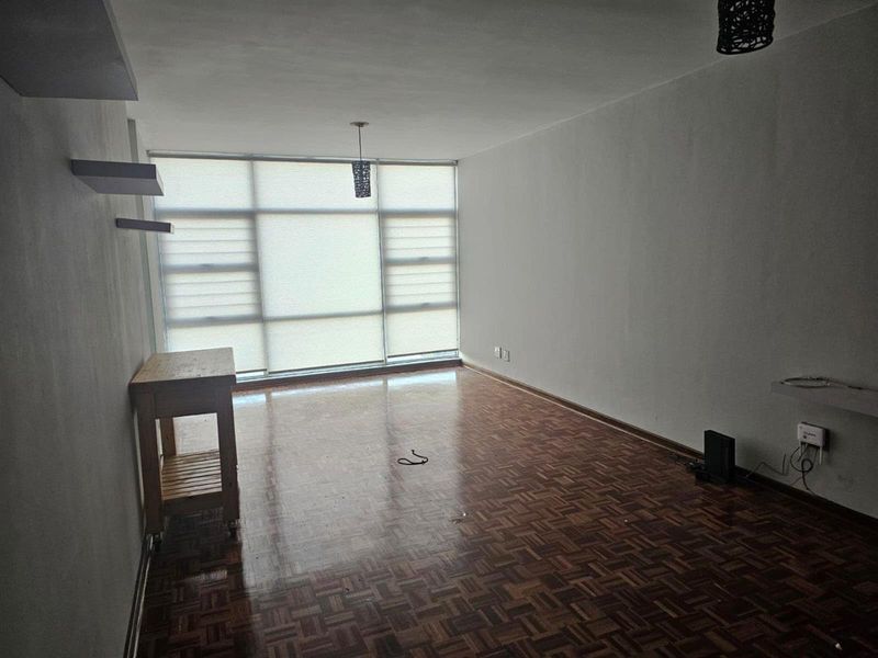 An empty living room featuring classic parquet flooring, large windows with roller blinds, and neutral walls, offering a blank canvas for personalization.