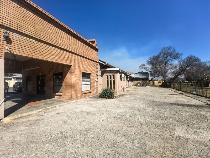Exterior view of the lodge featuring a face brick facade, covered entrance, and a large concrete driveway area with perimeter security fencing.