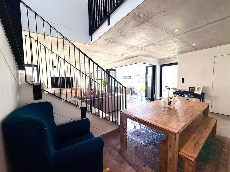 Modern open-plan living and dining area featuring an exposed concrete ceiling, sleek metal staircase railing, and large bi-fold doors opening to the outdoors.