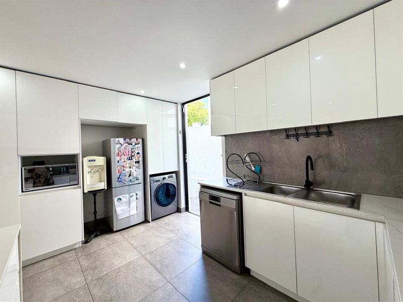Modern scullery featuring sleek white cabinetry, stone-look splashback, double sink, and integrated space for laundry appliances with direct outdoor access.