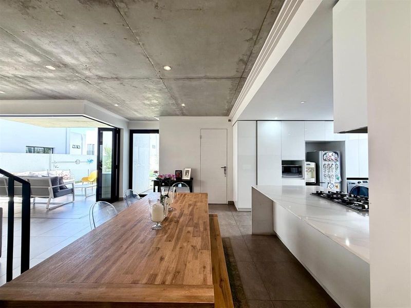 Modern open-plan living and kitchen area featuring an exposed concrete ceiling, sleek white cabinetry, stone island with gas hob, and seamless flow to the patio.