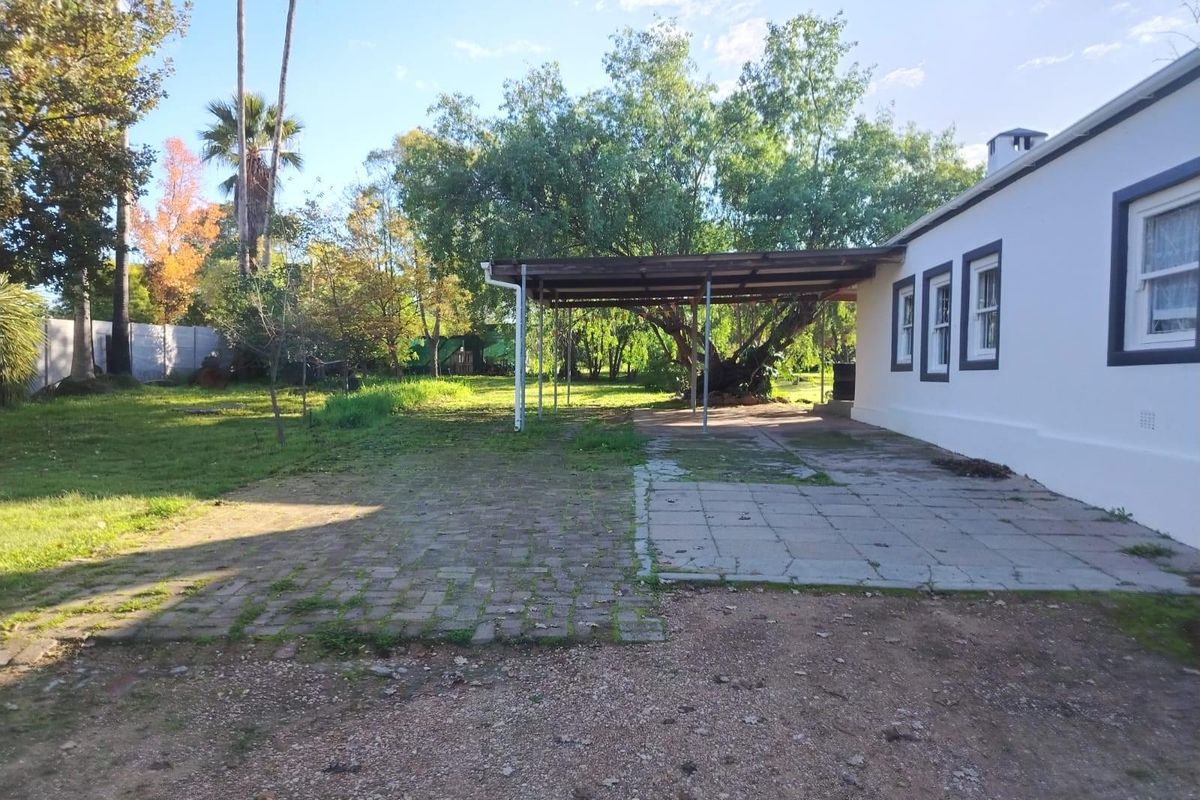 Rear exterior view featuring a paved patio area with a covered structure, overlooking a spacious garden with mature trees.