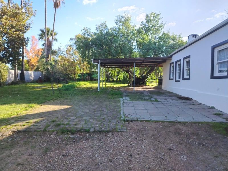 Rear exterior view featuring a paved patio area with a covered structure, overlooking a spacious garden with mature trees.