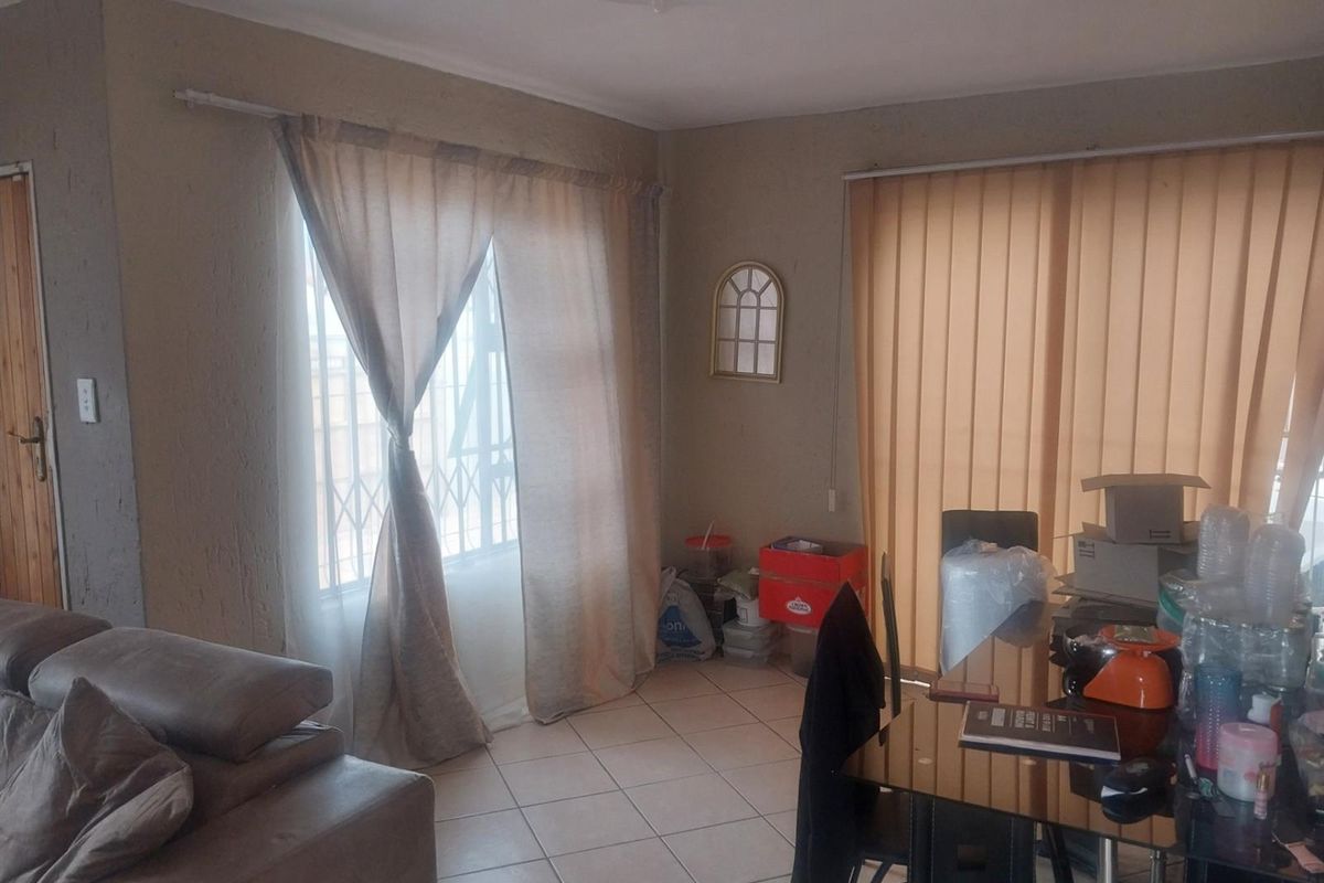 A living room area featuring tiled flooring, a decorative ceiling rose, and large windows fitted with security bars and vertical blinds.