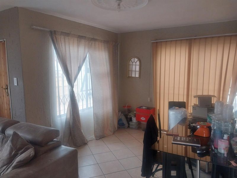 A living room area featuring tiled flooring, a decorative ceiling rose, and large windows fitted with security bars and vertical blinds.