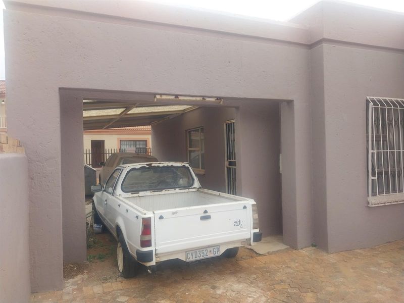 A covered carport area with paved flooring and security bars on the adjacent window, showing signs of wear and maintenance requirements.