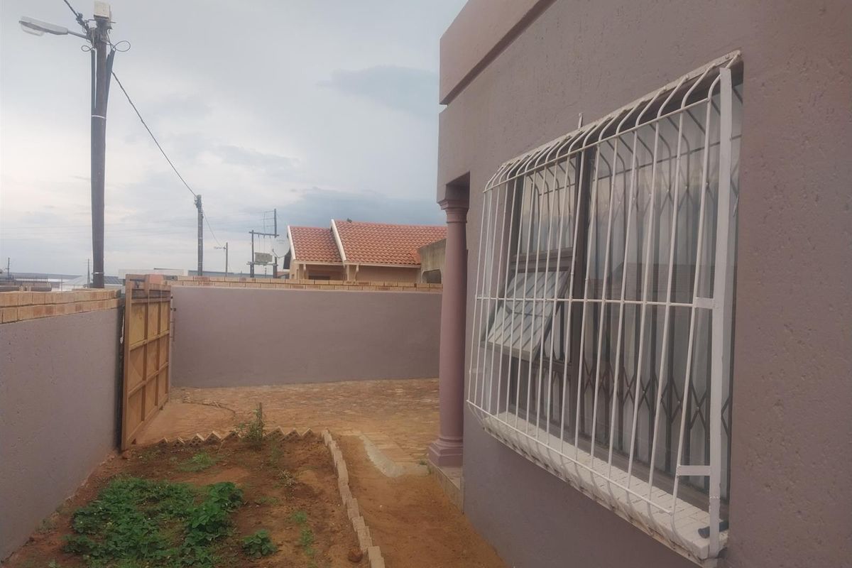 A view of the rear exterior area featuring a paved section, boundary wall, and a window fitted with security bars.