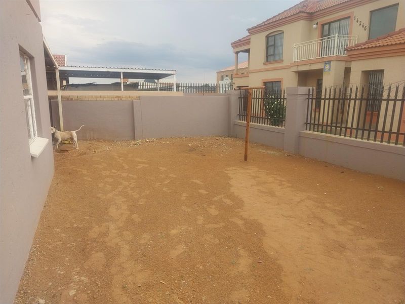A rear courtyard area featuring perimeter walls with security fencing and an adjacent carport structure, currently with an unlandscaped dirt surface.