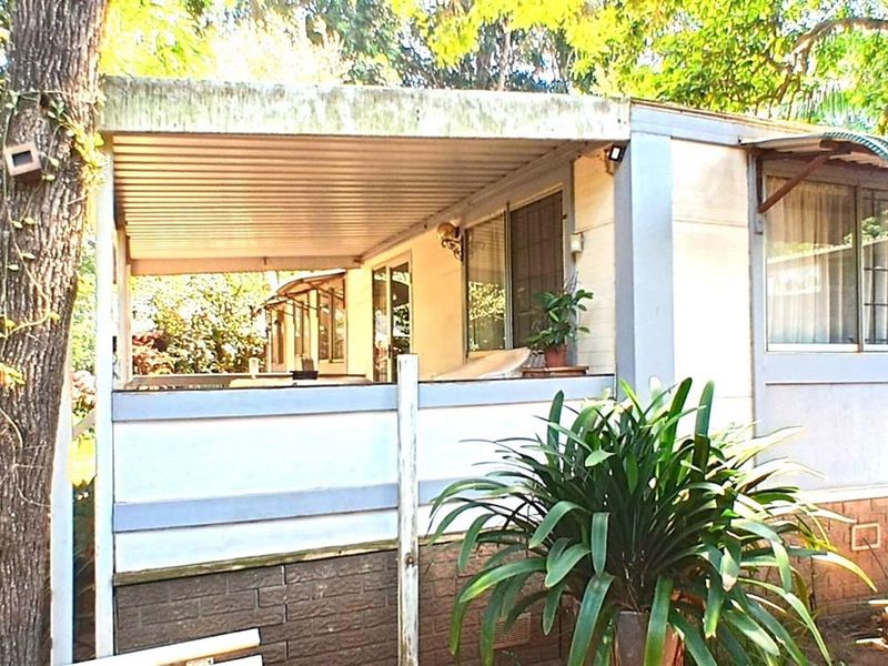 A covered outdoor patio area featuring a metal roof and brick base, set within a garden environment.