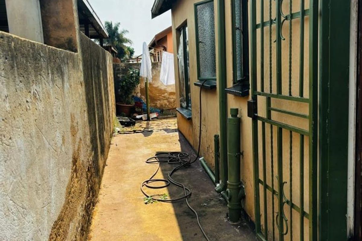 A narrow side exterior walkway featuring concrete paving, a security gate, and a boundary wall, showing signs of weathering and wear.