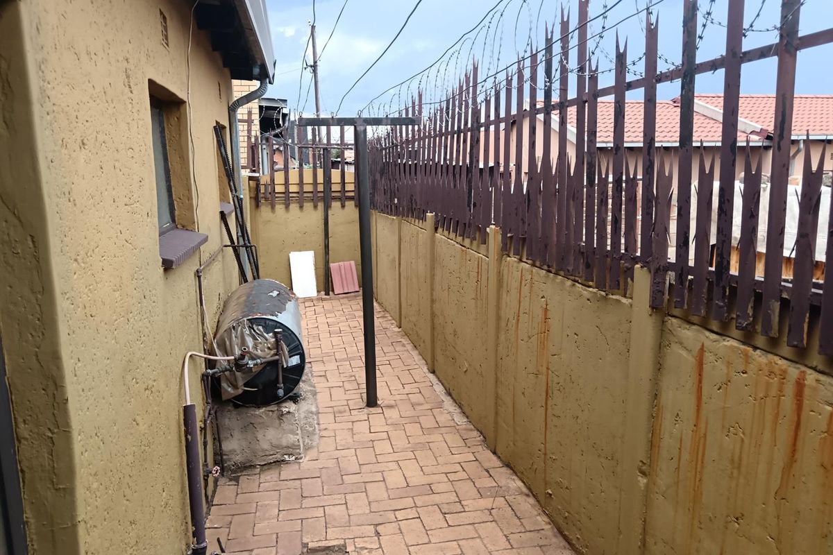 A narrow rear exterior side passage featuring paved flooring, a perimeter wall with security fencing, and an external water geyser unit.