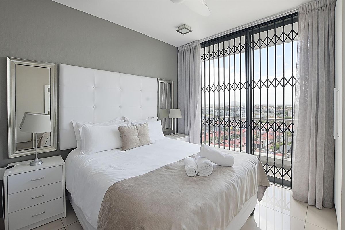 A well-maintained bedroom featuring tiled flooring, a large window with security gate, and access to a balcony with city views.