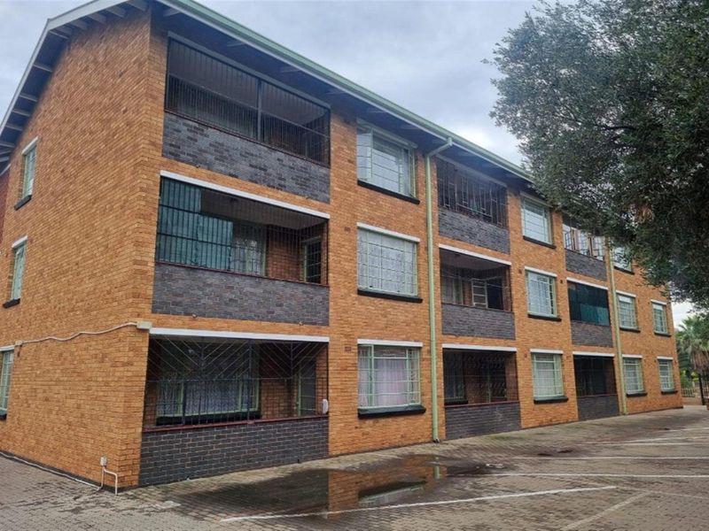 A multi-story residential apartment building featuring a brick facade, integrated balconies with security bars, and a paved parking area.