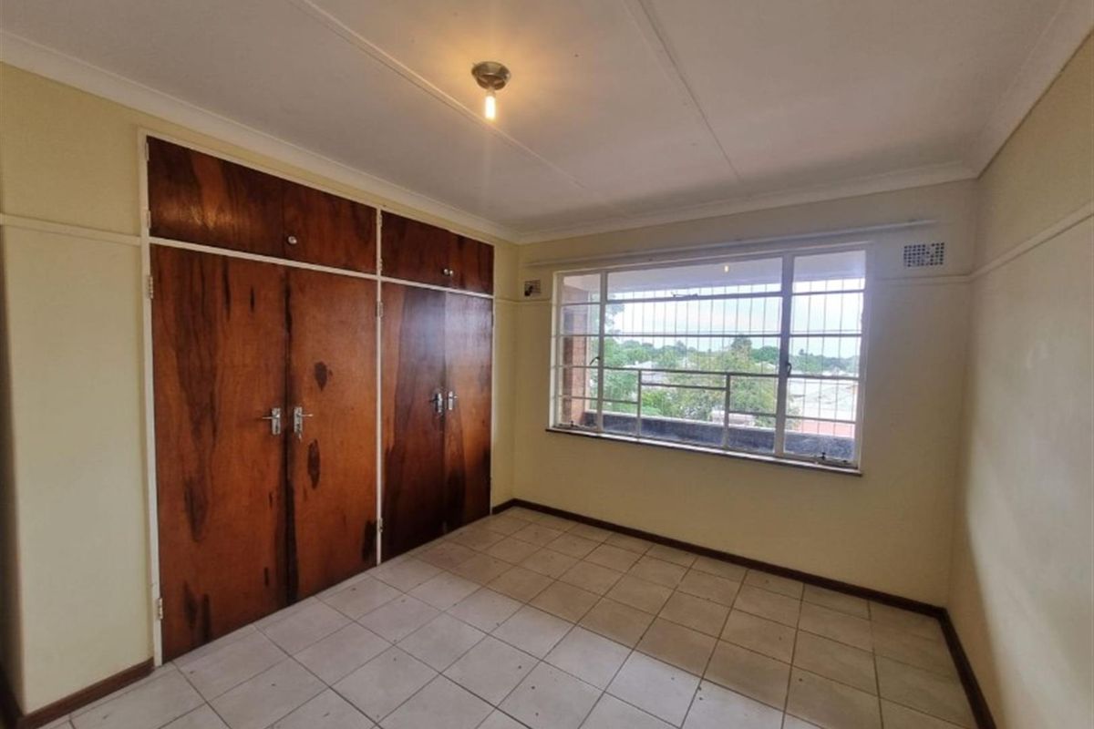 An empty bedroom featuring tiled flooring, built-in wooden cupboards, and a window with security bars, offering a functional space ready for personalization.