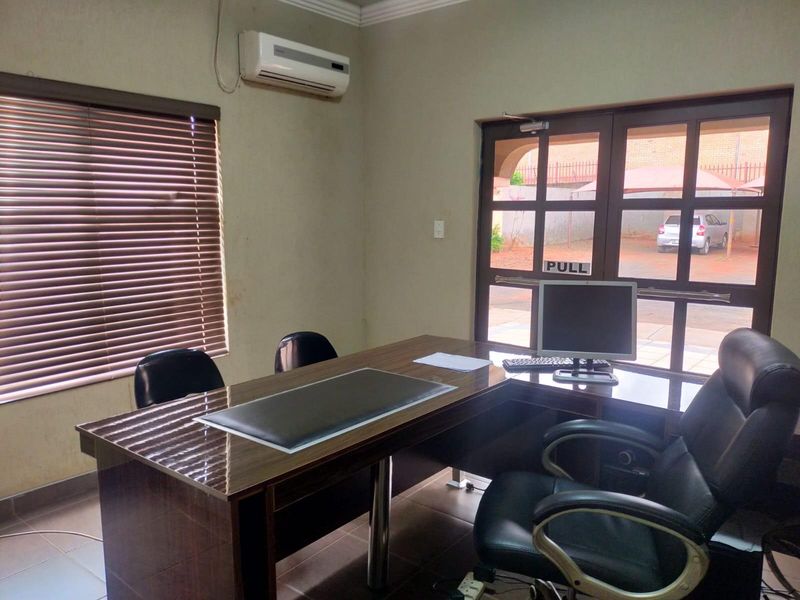 A functional office space featuring tiled flooring, a wall-mounted air conditioning unit, and large windows with venetian blinds providing natural light.