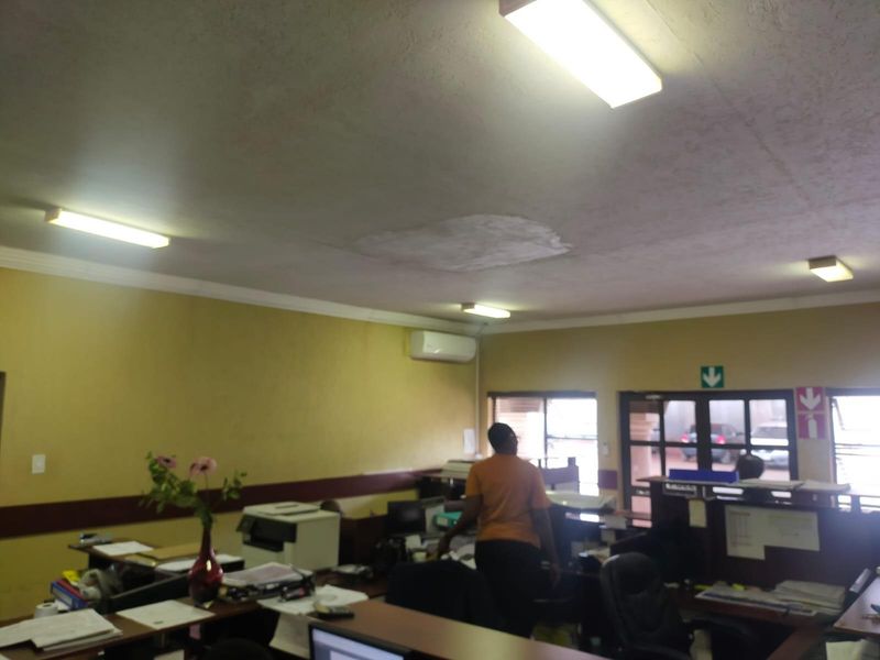 A functional office space featuring overhead fluorescent lighting, a wall-mounted air conditioning unit, and visible ceiling water damage requiring repair.