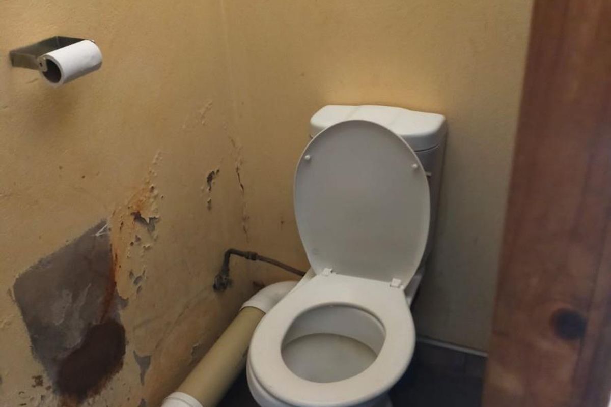 A small bathroom featuring a standard toilet and exposed plumbing, currently showing significant wear and damage to the wall finishes.