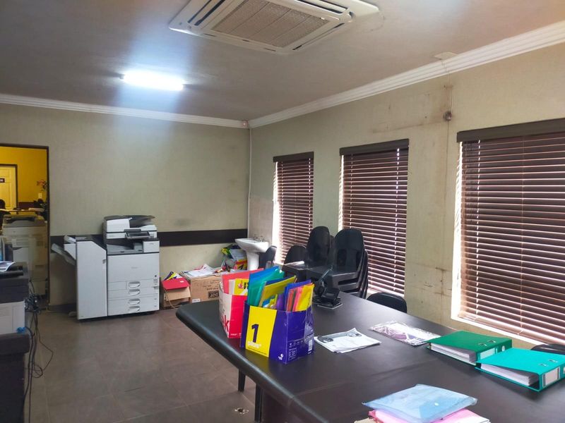 A functional office space featuring tiled flooring, ceiling-mounted air conditioning, and a small wall-mounted basin, with large windows fitted with venetian blinds.