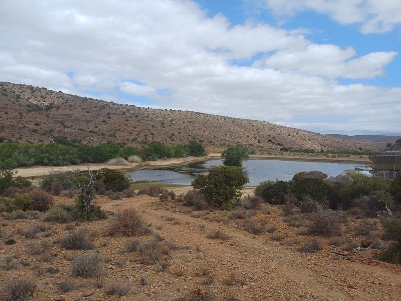 A scenic view of a natural farm dam surrounded by arid landscape and rolling hills under a partly cloudy sky.