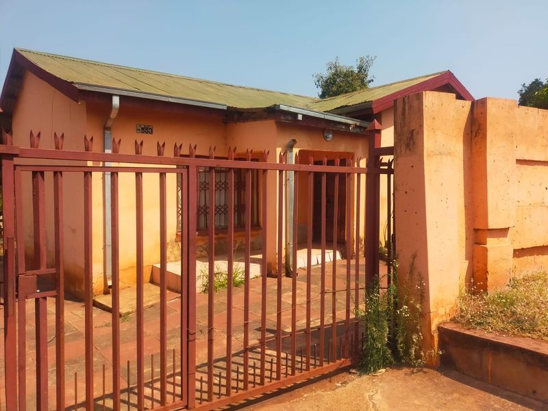 Front view of a single-story house featuring a metal security gate, perimeter wall, and paved entrance area requiring exterior maintenance.