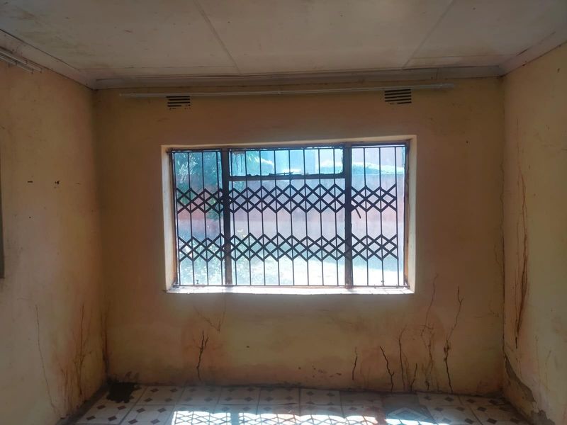 An empty bedroom featuring tiled flooring and a window with a security gate, showing significant signs of wall damage and wear.