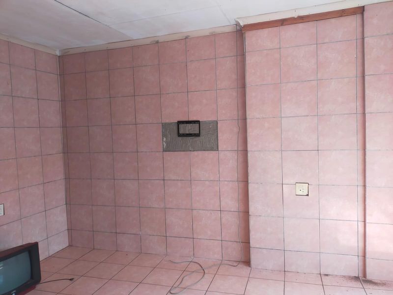 An empty living room space featuring dated pink ceramic wall and floor tiles, with a wall-mounted television bracket and exposed electrical wiring.