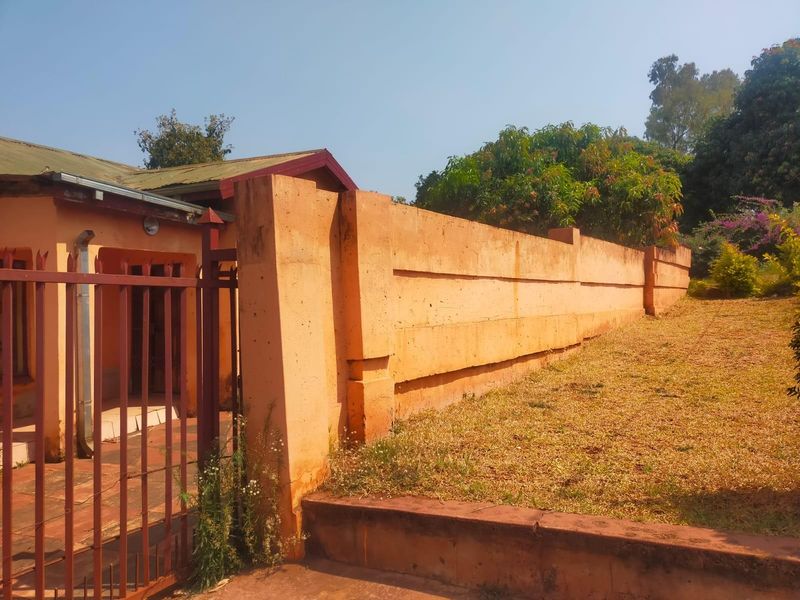 Front exterior view of the property featuring a long perimeter wall, metal gate, and paved entrance area with mature garden surroundings.