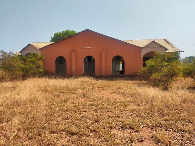Front view of a commercial building featuring a masonry facade with arched windows and a corrugated metal roof, set on an unmaintained plot.