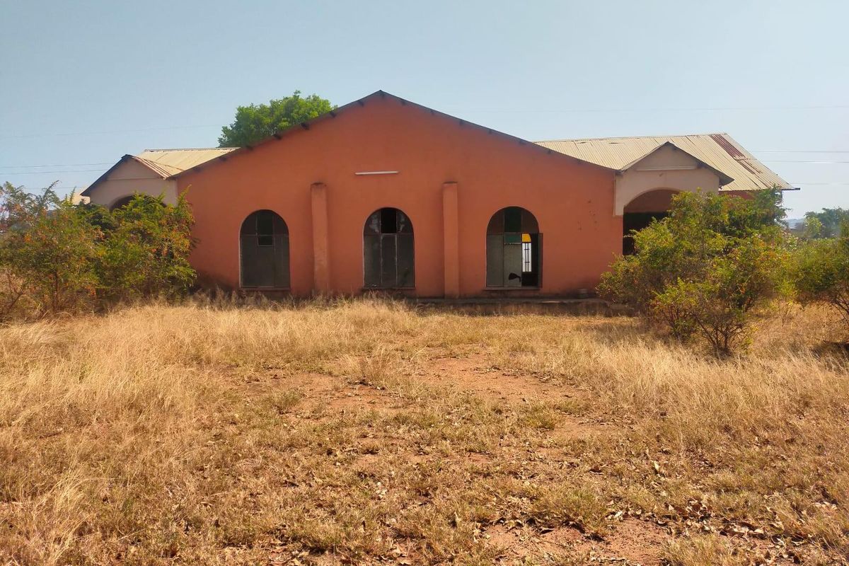 Front view of a commercial building featuring a masonry facade with arched windows and a corrugated metal roof, set on an unmaintained plot.