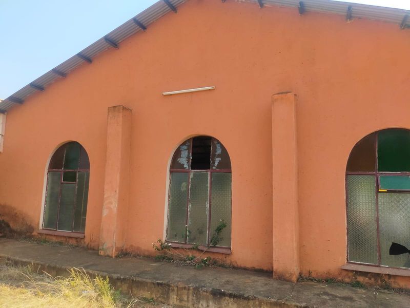 Exterior view of a commercial building featuring arched windows, a corrugated metal roof, and a concrete walkway, showing signs of significant wear and damage.